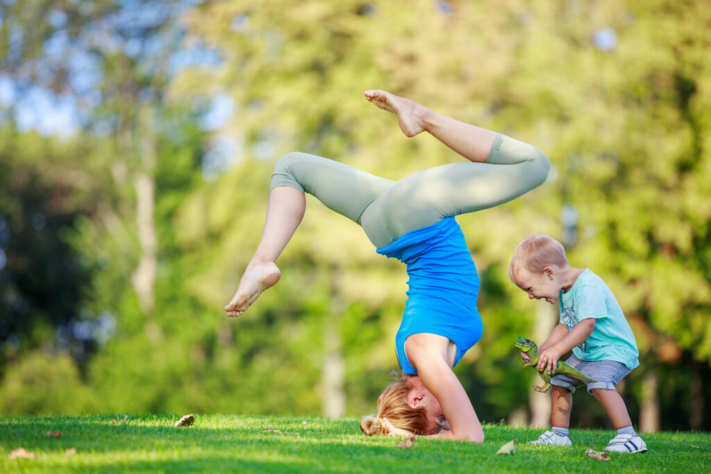 Young woman working out outdoors, standing on head, little son playing beside her